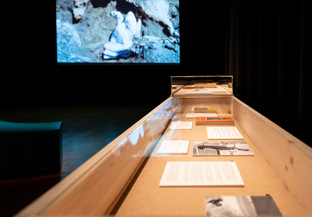 In the foregrund is a wooden display box with perspex covers, housing a collection of printed texts, books and photographic material situated in a dark room. On the left side, faintly spotlit is a curved seat, in front of a large projection screen that is slightly out of focus. It appears to have an African woman with a face covering, wearing a white dress is lying in a mountainous rock surface. 