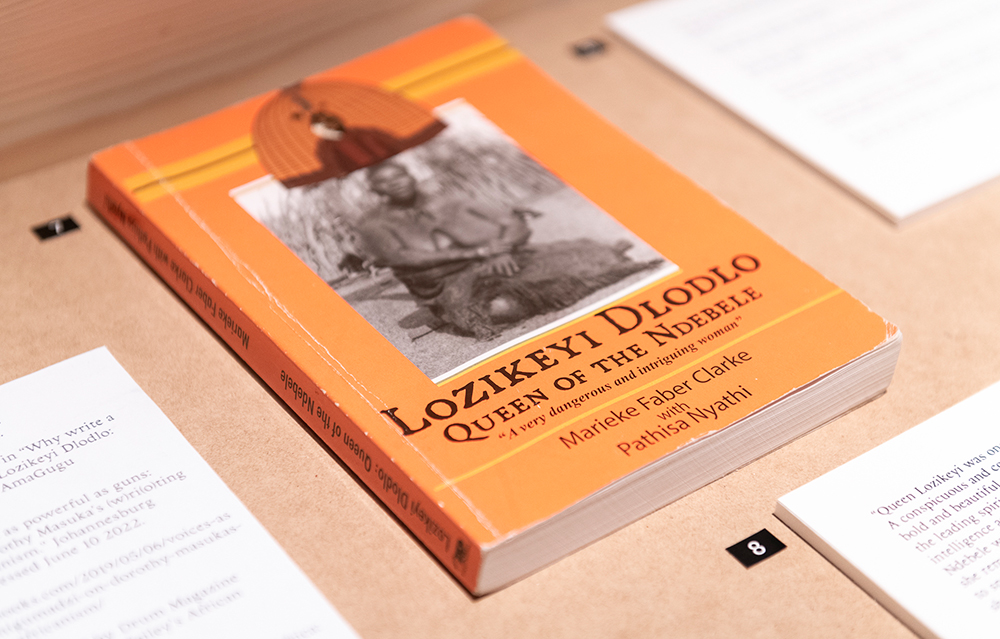 A bright orange book with a monochrome image of an African woman on its cover reads "Lozikeyi Dlodlo, Queen of the Ndebele". It sits on a light brown table, with printed material adjacent that has text that is out of focus.