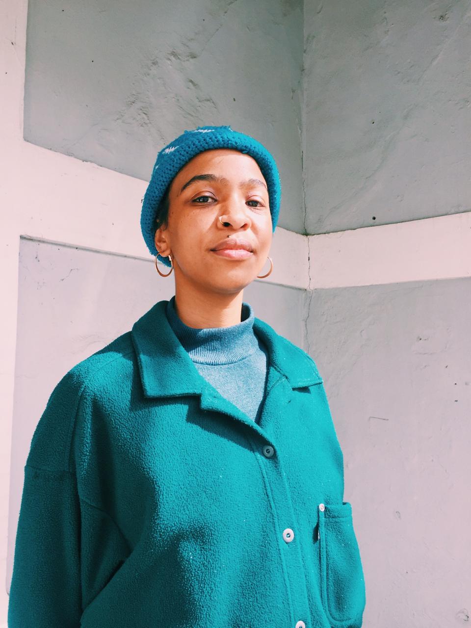 An African women wearing hoop earrings, a blue beanie, blue turtle neck and teal wool cardigan is standing tall in front of a concrete painted wall.