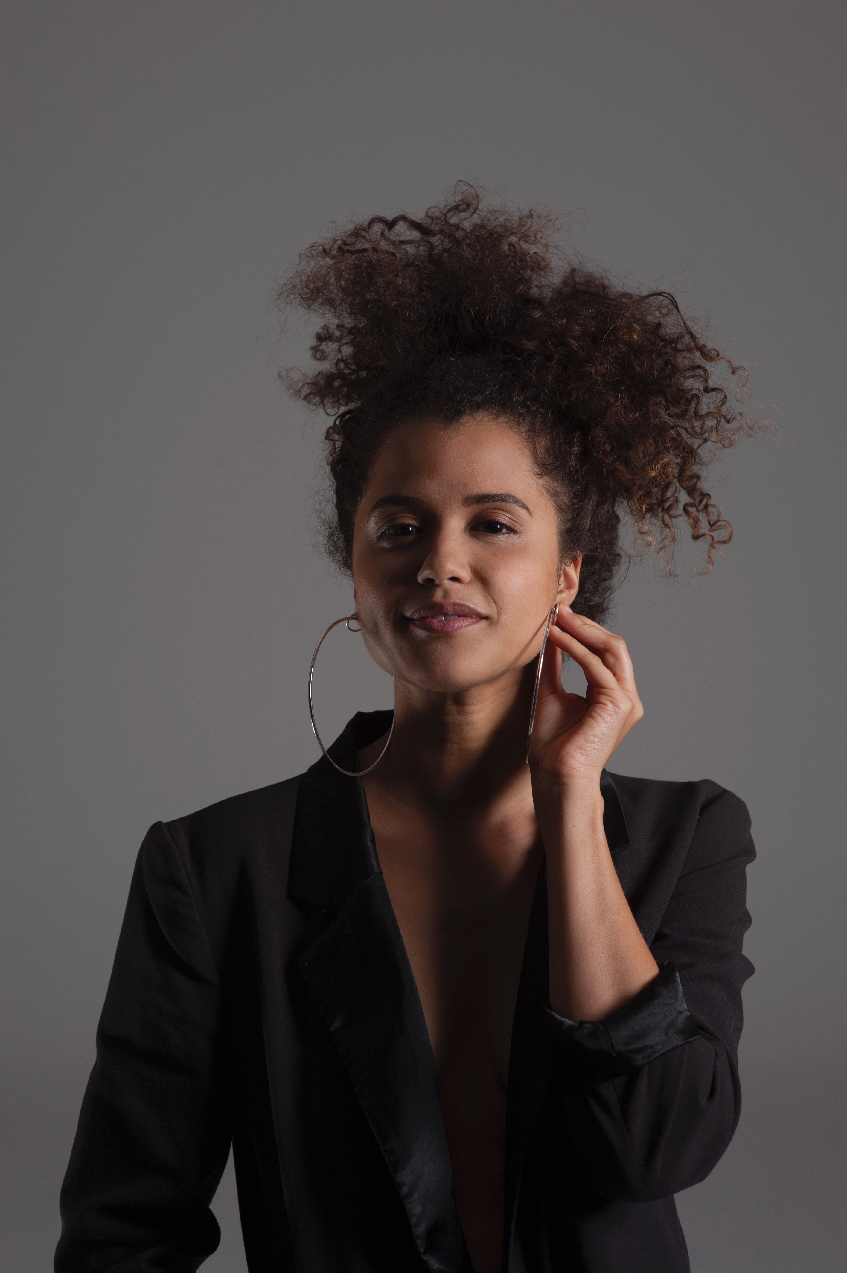 From the waist up in front of a grey background is an African woman with light complexion and big curly hair tied up. She is holding her very large hoop earrings and wearing a black blazer suit jacket.