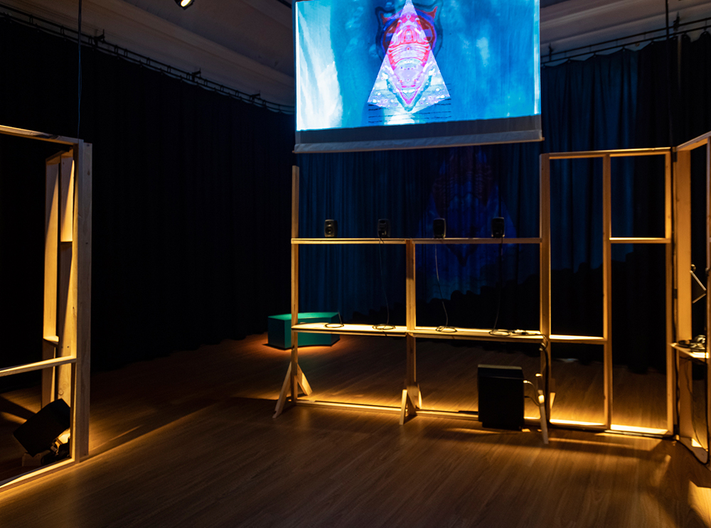 A dark room bordered with black curtains and wooden floorboards has installations of pine timber frame structures with speakers mounted within the structure. A video projection hangs above with an abstract drawing in red hues and background with shades of blue.